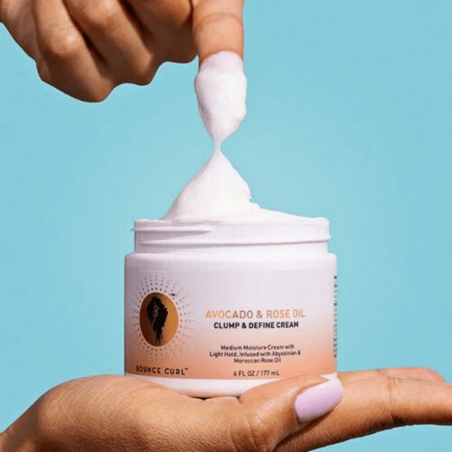 Hand entnimmt weiße Creme aus einem Tiegel „Avocado & Rose Oil Clump & Define Cream“ vor blauem Hintergrund.