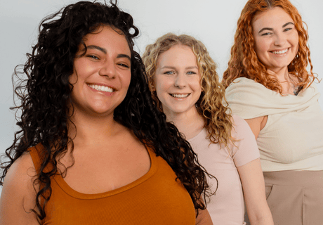 Zur Lockenberatung – Drei Frauen mit lockigem Haar lächeln in die Kamera, Bild führt zur Buchung eines Beratungstermins.