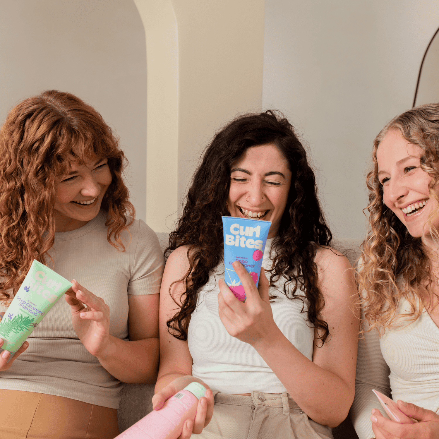 Zum Lockenquiz – Drei Frauen mit lockigem Haar lachen und halten Haarpflegeprodukte, Bild führt zum Lockenquiz.